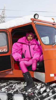 a woman sitting in the door of an orange truck on snow covered ground with skis