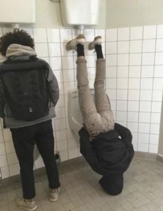 two people standing in a bathroom while one person is upside down on the urinal