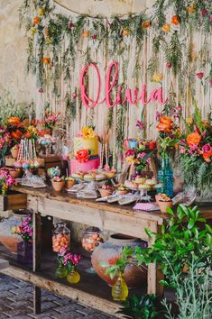 a table topped with lots of cakes and flowers next to a wall covered in greenery