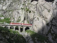 a red train traveling over a bridge on top of a rocky mountain side next to a cliff