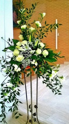 a tall metal vase with white flowers and greenery in it sitting on a stand