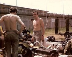 two men standing in front of an old race car and another man with no shirt on