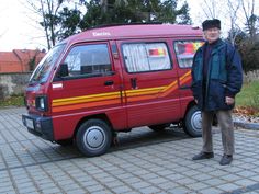 a man standing next to a red van on a brick road with trees in the background