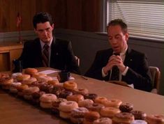 two men sitting at a table with donuts in front of them on the table