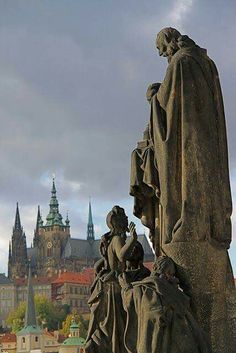 the statue is in front of an old city with many spires and buildings behind it