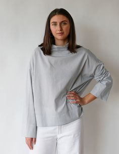 a woman standing in front of a white wall wearing a gray sweater and white pants