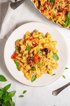 a plate of pasta with tomatoes and basil next to a casserole dish on a white table