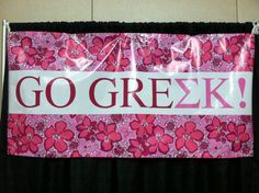 a sign that says go greek with flowers on it hanging from a black curtain in front of a wall