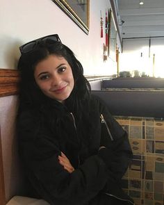 a woman with long black hair and sunglasses sitting on a bench next to a wall