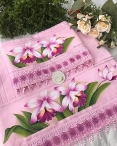 two pink towels with purple flowers on them next to some white roses and greenery