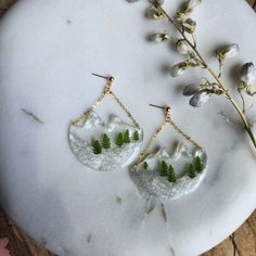 two earrings with green leaves hanging from them on a white marble table next to dried flowers