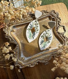 two white and gold earrings sitting on top of a wooden table next to dried flowers