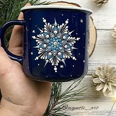 a hand holding a blue coffee mug with snowflakes on it and pine cones in the background