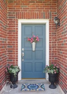 two planters with flowers are on the front steps of a brick building, next to a blue door