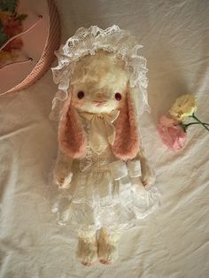 a stuffed rabbit wearing a white dress and bonnet with flowers on the bed next to it