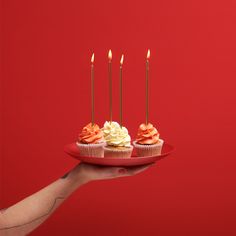 a hand holding a plate with cupcakes and candles on it in front of a red background