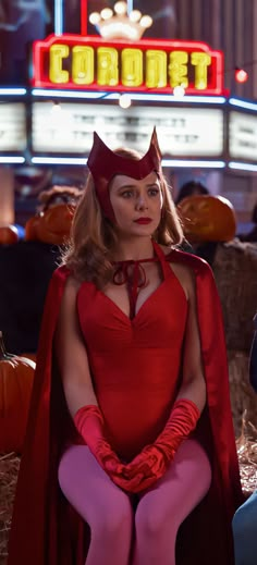 a woman in a red dress and catwoman costume sitting on hay with a neon sign behind her