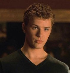 a young man with curly hair wearing a black v - neck shirt and looking at the camera