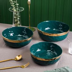 three teal bowls with gold rims on a table next to two forks and spoons