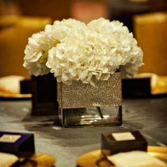 white flowers are in a square vase on a table with gold plates and napkins