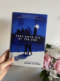 the book they both die at the end by adam silvera is held up in front of flowers