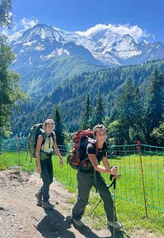 two people with backpacks walking up a trail in front of mountains and trees on a sunny day