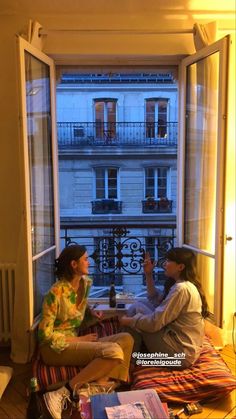 two women sitting on a bed in front of an open window looking at each other