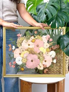 a woman holding up a framed painting with flowers in the center and greenery behind it