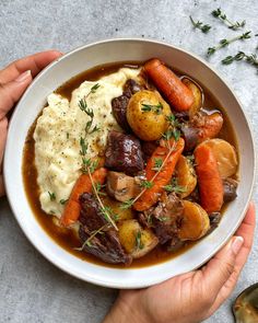 two hands holding a bowl of stew and mashed potatoes with carrots on the side