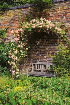 a wooden bench sitting in the middle of a lush green garden next to a brick wall