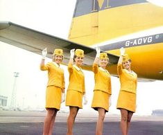 four women in yellow uniforms standing next to an airplane on the tarmac with their hands up