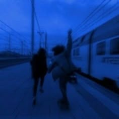 two people are walking down the sidewalk near a train track at night, one person is holding his hand up