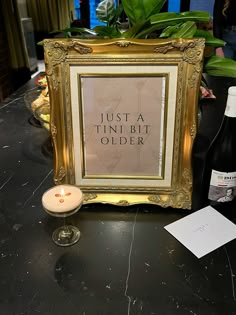 a table topped with a bottle of wine next to a framed sign that says just a tinni bit older