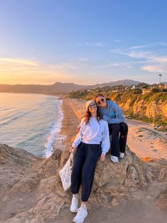 two people sitting on top of a rock next to the ocean