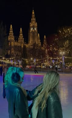 two people standing on an ice rink at night with lights and buildings in the background