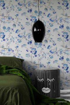 A cozy bedroom corner featuring a botanical chinoiserie wallpaper with blue flowers and vines. A green bedspread and matching throw drape over the bed, while a black cylindrical stool with a face illustration stands beside it. A glossy black pendant lamp hangs overhead. Blue Botanical Wallpaper, White Blue Wallpaper, White And Blue Wallpaper, Wallpaper Chinoiserie, Wall Taps, Chinoiserie Wallpaper