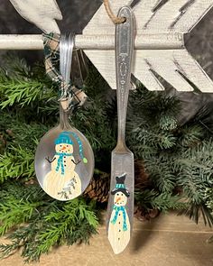 two spoons with snowmen on them hanging from a christmas ornament next to evergreen branches