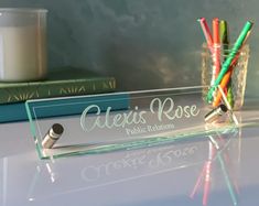 a clear glass desk sign with colored pencils in it next to a stack of books