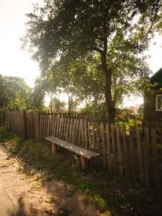 a wooden bench sitting next to a tree on the side of a road in front of a fence