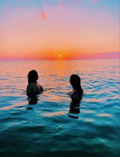 two women are sitting in the water watching the sun go down over the ocean with their backs to each other