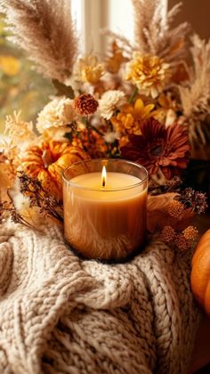 a candle sitting on top of a blanket next to flowers and pumpkins in front of a window