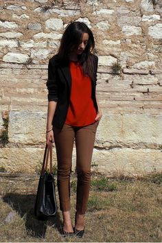 a woman standing in front of a stone wall holding a black purse and wearing brown pants