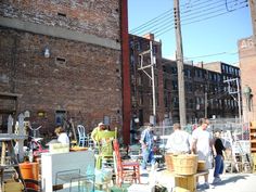 several people are standing outside near many chairs and other items on the sidewalk in front of an old brick building