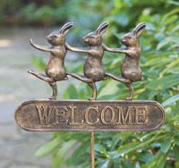 a metal welcome sign with three rabbits on it and the words welcome are in front of some bushes