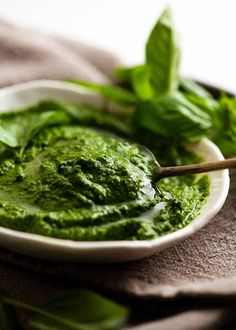 a white bowl filled with green pesto on top of a cloth next to a wooden spoon