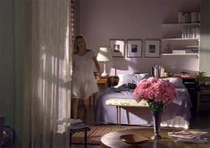 a woman standing next to a bed in a room with pink flowers on the table