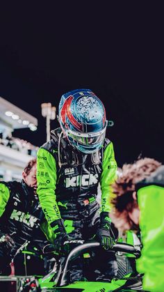 a person in a racing suit and helmet sitting on a motorcycle with other people behind him