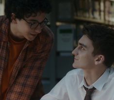 two young men sitting next to each other in front of a bookshelf and looking at each other