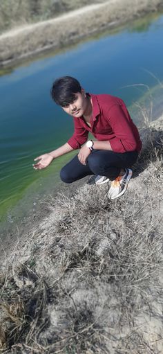 a young man crouches on the edge of a body of water while holding his hand out