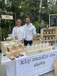 two people standing behind a table with candles and jars on it at an outdoor market